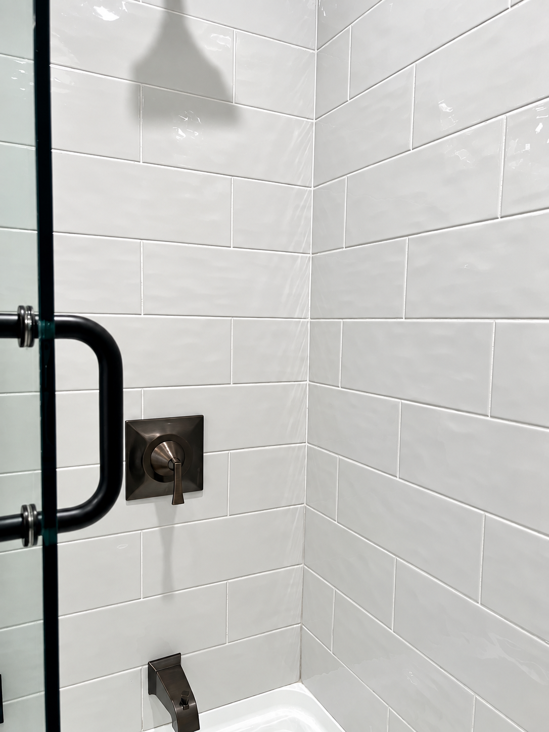 White tiled shower corner with bronze fixtures and a glass door